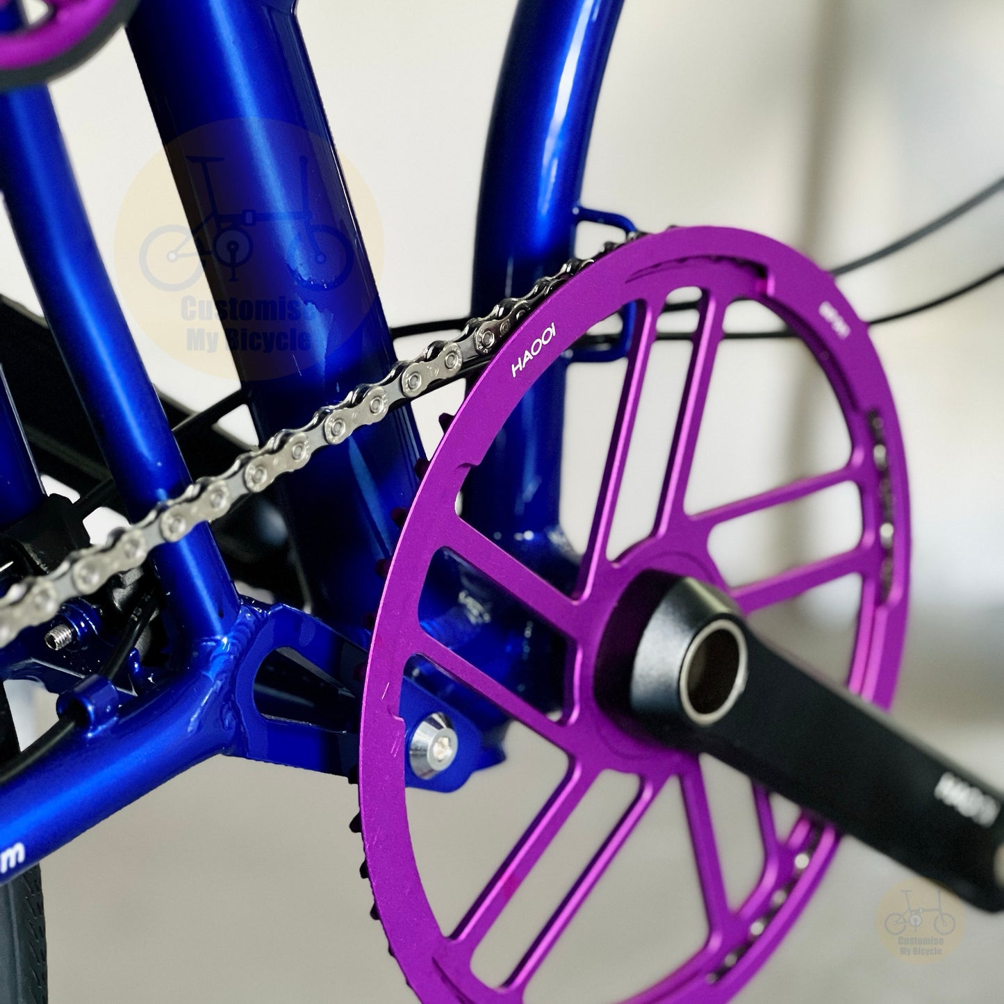 Close-up of a bicycle crankset with a bright purple chainring and black crank arm, attached to a metallic blue frame. A silver chain is visible, and small white branding HAOOI appears on the chainring and crank arm.