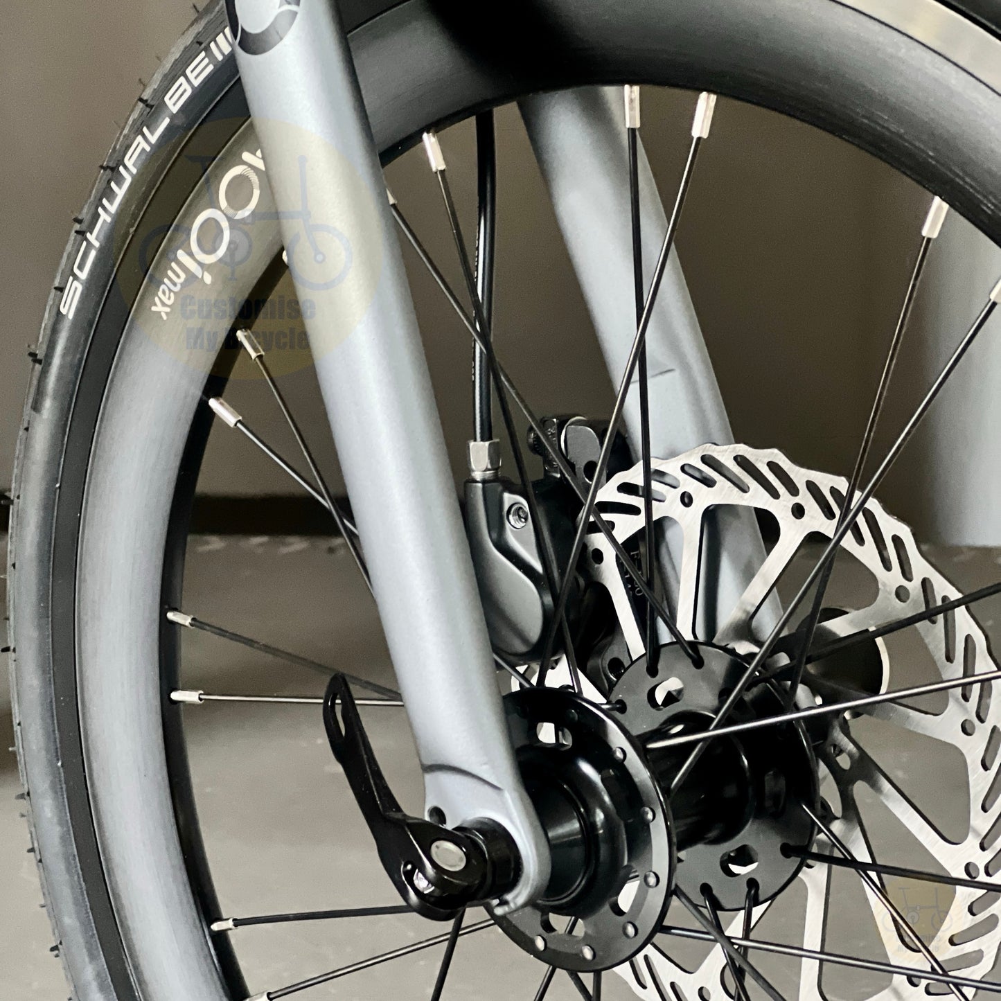 Close-up of a bicycle wheel and front fork showing a silver disc brake rotor, black spokes, and part of a black tire with white branding text.