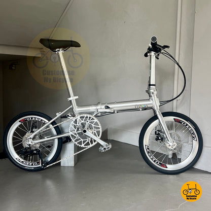Full side view of Fnhon Zephyr 18-inch folding bike in polished silver, displayed on wooden deck with city skyline at sunset.
